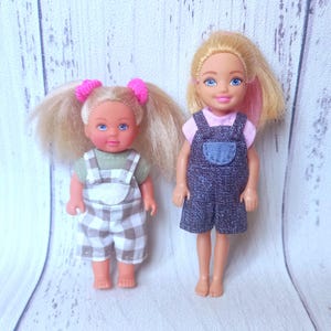 May include: Two small dolls with blonde hair and blue eyes. One doll wears a green shirt and plaid overalls. The other doll wears a pink shirt under a denim jumper. Both dolls are standing upright.