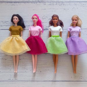 May include: Four fashion dolls wearing colorful outfits. Each doll has a different hairstyle and skin tone. The dolls are dressed in short-sleeved tops and tulle skirts in shades of yellow, pink, green, and purple.