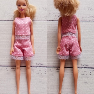 May include: A doll in a pink polka dot pyjama set. The top and shorts are trimmed with white lace. The doll has blonde hair and is positioned against a light-coloured background.