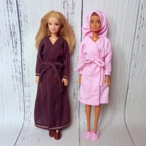 May include: Two dolls wearing robes. One doll is in a dark burgundy robe with a matching belt. The other doll is in a light pink robe with a hood and a matching belt. Both dolls are standing on a light wood surface.