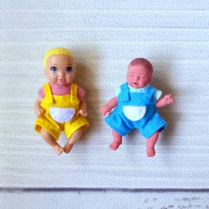 Pode incluir: Duas pequenas bonecas. Uma boneca tem macacão amarelo, camisa rosa e cabelo loiro. A outra boneca tem macacão azul e camisa branca. Ambas as bonecas estão sentadas.