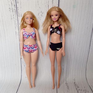 May include: Two blonde dolls wearing swimwear. One doll is wearing a two-piece swimsuit with a white, red, and blue pattern. The other doll is wearing a black two-piece swimsuit with a floral pattern.