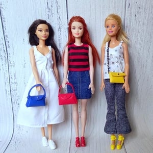 May include: Three fashion dolls are displayed, each showcasing different outfits. One doll wears a white dress and carries a blue handbag. Another doll is dressed in a red and black striped top and denim skirt, holding a red handbag. The third doll has a white floral top, gray pants, and a yellow purse.