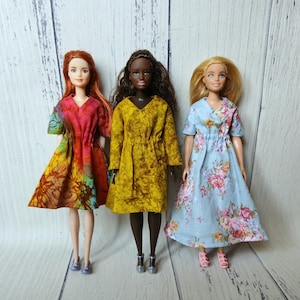 May include: Three fashion dolls standing side by side. The left doll has red hair and wears a short-sleeved dress with a multicolored tie-dye pattern. The center doll has dark skin and wears a mustard yellow dress. The right doll has blonde hair and wears a light blue dress with a pink floral print.