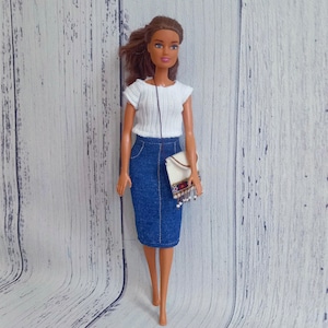 May include: A doll wearing a white knit top, a denim pencil skirt, and carrying a small white purse with decorative trim. The doll has brown hair and is posed against a light wood-grain background.