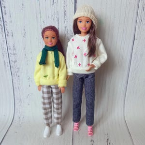 Puede incluir: Dos muñecas vestidas con atuendos de invierno. Una muñeca lleva un suéter amarillo, pantalones a cuadros, una bufanda verde y zapatillas blancas. La otra muñeca lleva un suéter blanco con detalles florales rosas, pantalones oscuros, un gorro de punto y zapatos rosas.