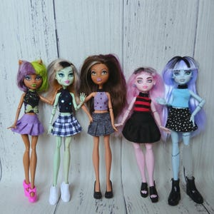 May include: Five fashion dolls standing in a row. The first has purple and green hair, a black butterfly top, and a purple skirt. The second has pale green skin, a black top, and a plaid skirt. The third has brown hair, a purple top, and a grey skirt. The fourth has pink hair, a red and black striped top, and a black skirt. The fifth has blue hair, a light blue top, and a black and white polka dot skirt.