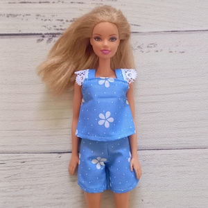 Könnte beinhalten: Eine Puppe in einem blauen Oberteil und Shorts-Set. Das Outfit hat ein weißes Blumenmuster und weiße Tupfen. Das Oberteil hat eine weiße Spitzentrimmung an den Trägern. Die Puppe hat blondes Haar.