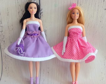 Könnte beinhalten: Zwei Modepuppen in eleganten Kleidern. Eine Puppe trägt ein lila Kleid mit weißem Besatz, Handschuhen und einer Tiara. Die andere Puppe trägt ein rosa Kleid mit weißen Punkten, weißem Besatz und einer Schleife. Beide Puppen haben passende Schuhe.