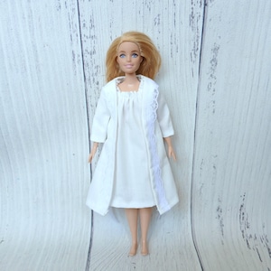 May include: A doll with blonde hair wearing a white sleeveless dress and a matching white open-front robe. The robe features delicate white lace trim along the front edges. The doll has blue eyes and is standing in an upright position.