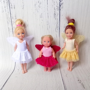 May include: Three small fairy dolls with blonde and brunette hair, wearing dresses and wings. One doll wears a white dress with white wings, another a pink dress with red wings, and the third a yellow dress with yellow wings.