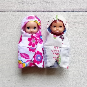 May include: Two small baby dolls swaddled in floral blankets. One doll is wrapped in a white blanket with pink and blue flowers, the other in a white blanket with red and pink floral patterns. Both dolls have hooded blankets.
