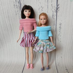 May include: Two dolls standing side by side. The doll on the left has dark hair, a pink off-the-shoulder top, and a red and white striped skirt. The doll on the right has light hair, a light blue off-the-shoulder top, and a floral print skirt.