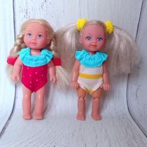 May include: Two small dolls with blonde hair and blue eyes. One doll wears a red swimsuit with white polka dots and a turquoise ruffled collar. The other doll wears a white swimsuit with yellow stripes and a turquoise ruffled collar.