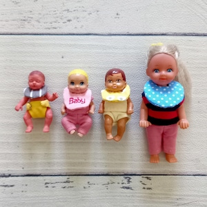 May include: Four small baby dolls. One doll wears a yellow diaper and a gray bib. Another doll wears pink pants and a bib that says "Baby". The third doll wears a yellow bib and a yellow onesie. The last doll wears pink pants, a blue and white polka dot bib, and a black and red striped shirt.