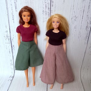 May include: Two dolls wearing handmade doll clothes. One doll is wearing a burgundy t-shirt and green culottes. The other doll is wearing a black t-shirt and mauve culottes. The dolls are standing in front of a light wood background.