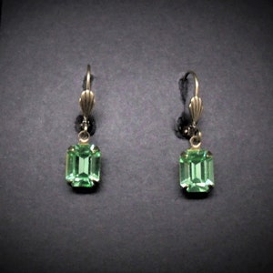 May include: A pair of vintage-style drop earrings. Each earring features a rectangular, light green gemstone set in a gold-toned metal frame. The earrings have a lever-back closure and a small black bead detail.