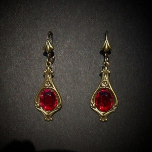 Op de afbeelding: Een paar gouden hangende oorbellen met rode glazen stenen. De oorbellen hebben een traanvorm en ingewikkelde bloemmotieven.