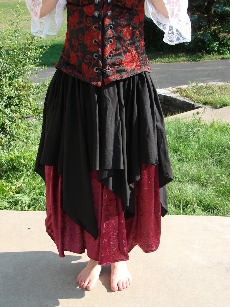 May include: A black and red skirt with a layered design. The top layer is a black skirt with a ruffled hem. The bottom layer is a burgundy velvet skirt. The skirt is worn with a black corset with red floral embroidery.