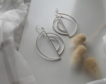 Puede incluir: Un par de pendientes geométricos plateados con un diseño de medio círculo. Los pendientes están hechos de metal fino y tienen un estilo moderno y minimalista.