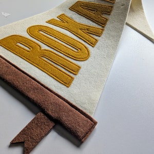 May include: Cream-colored felt pennant with the word "ROKA" in gold lettering. The pennant has a brown border and a brown ribbon. The pennant is likely a decorative item.