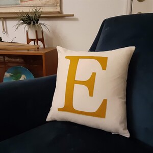 May include: A white throw pillow with a large, mustard yellow letter "E" in the center. The pillow is sitting on a dark blue armchair.