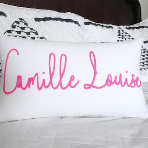 May include: A white rectangular pillow with the name "Camille Louise" in bright pink cursive lettering. The pillow is on a bed with white bedding and decorative pillows with black and white geometric patterns.