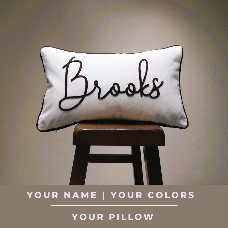 Puede incluir: Almohada blanca con un nombre bordado en negro "Brooks" en ella. La almohada est&aacute; sentada en un taburete de madera.