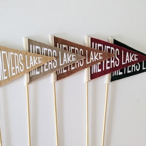May include: Five felt pennant flags with wooden sticks. Each flag is a different color: tan, brown, orange, red, and black. The flags all say "Meyers Lake" in white lettering.