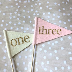 May include: Two triangular felt flags on wooden sticks. One flag is pale yellow with the word "one" in gold glitter. The other is pink with the word "three" in gold glitter. The background is a beige fabric with white polka dots.