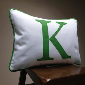 May include: White pillow with a green felt letter K in the center. The pillow has a green border and is sitting on a wooden chair.