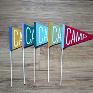 May include: Five colorful felt pennant flags on wooden sticks. The flags are in shades of yellow, purple, teal, light blue, and red. The flags spell out "CA CA CA CA CAMP" in white letters. The background is a wooden surface.