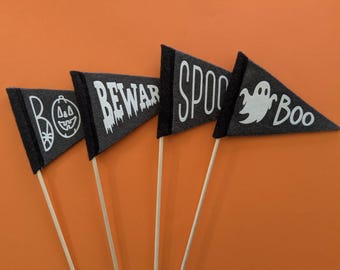 May include: Four gray felt pennant flags on wooden sticks with Halloween-themed text and graphics. The flags read "BOO" with a jack-o'-lantern, "BEWARE", "SPOOK", and "Boo" with a ghost. The flags have black trim and are set against an orange background.