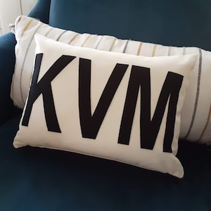 May include: A white pillow with the letters "KVM" in black felt on the front. The pillow is on a teal couch.
