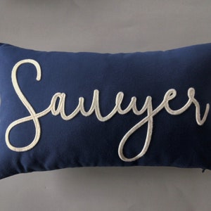 May include: A navy blue pillow with the name "Sawyer" embroidered in white thread.