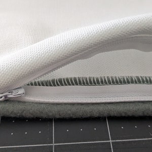 Puede incluir: Una funda de almohada blanca con cierre de cremallera. La cremallera est&aacute; abierta y el interior de la funda de almohada es visible. La funda de almohada est&aacute; sobre una superficie gris.