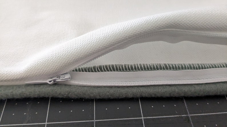 Puede incluir: Una funda de almohada blanca con cierre de cremallera. La cremallera est&aacute; abierta y el interior de la funda de almohada es visible. La funda de almohada est&aacute; sobre una superficie gris.