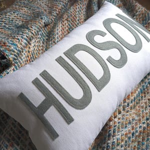 May include: White rectangular pillow with the name "HUDSON" in gray letters. The pillow is resting on a patterned blanket with blue, orange, and brown hues. The pillow is a decorative home accent.