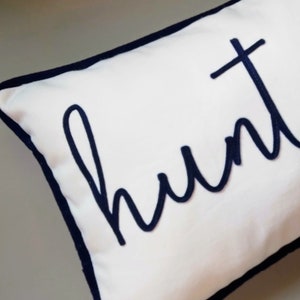 Puede incluir: Almohada blanca con borde azul marino y la palabra "hunt" bordada en cursiva azul marino.