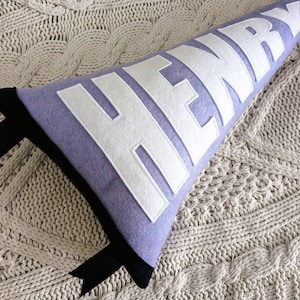 May include: A purple felt pillow with the name "HENRY" in white felt letters. The pillow has black trim and is on a cream knitted surface.