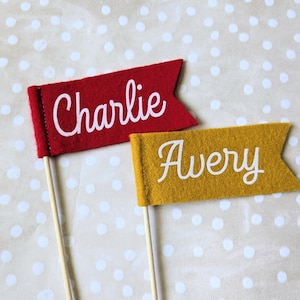May include: Two felt flag decorations on wooden sticks. One is red with the name "Charlie" in white script, and the other is yellow with the name "Avery" in white script. The background is a light beige with white polka dots.