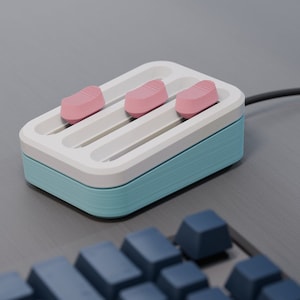 May include: A three-button device with a light blue base, white top, and three pink buttons. The device is connected to a black cable and sits on a gray surface. Blue keyboard keys are visible in the foreground.