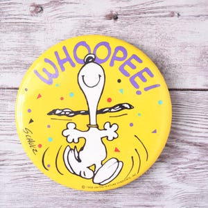 May include: A round, yellow Snoopy button with the word "WHOOPEE!" in purple. Snoopy is depicted in white with black outlines, celebrating. The button has colorful confetti and the text "Schulz".