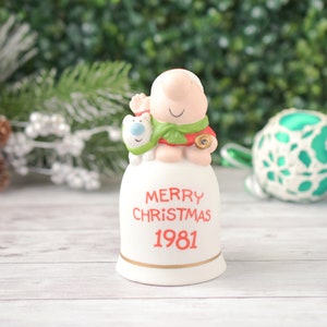 May include: A vintage ceramic Christmas bell figurine. The bell features a smiling character with a polar bear, wearing a green scarf and red outfit. The bell reads "MERRY CHRISTMAS 1981" in red lettering with a gold trim.