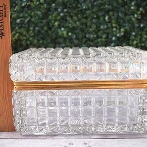 K&ouml;nnte beinhalten: Eine rechteckige, transparente Glasschmuckschatulle mit einem Rastermuster und goldfarbenem Rand. Die Box ist etwa 15 cm lang. Deckel und Seiten weisen ein sich wiederholendes quadratisches Design auf. Die Box steht auf einer wei&szlig;en Oberfl&auml;che.