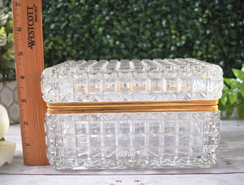 K&ouml;nnte beinhalten: Eine rechteckige, transparente Glasschmuckschatulle mit einem Rastermuster und goldfarbenem Rand. Die Box ist etwa 15 cm lang. Deckel und Seiten weisen ein sich wiederholendes quadratisches Design auf. Die Box steht auf einer wei&szlig;en Oberfl&auml;che.