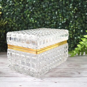 K&ouml;nnte beinhalten: Eine transparente, rechteckige Glasbox mit einem strukturierten, gitterartigen Muster. Die Box hat ein goldfarbenes Metallband um die Mitte. Der Deckel ist ebenfalls strukturiert und passt gut oben drauf. Die Box ist wahrscheinlich f&uuml;r Schmuck oder kleine Gegenst&auml;nde.