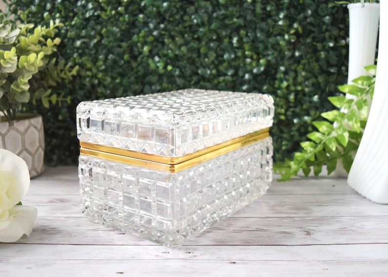 K&ouml;nnte beinhalten: Eine transparente, rechteckige Glasbox mit einem strukturierten, gitterartigen Muster. Die Box hat ein goldfarbenes Metallband um die Mitte. Der Deckel ist ebenfalls strukturiert und passt gut oben drauf. Die Box ist wahrscheinlich f&uuml;r Schmuck oder kleine Gegenst&auml;nde.