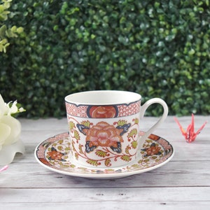 May include: A white teacup and saucer set with a floral design in orange, blue, and green. The cup and saucer have a decorative border. The set is on a white wooden surface.