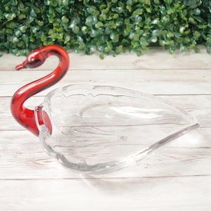 May include: A clear glass swan-shaped dish with a red glass swan head and neck. The dish has a leaf-like shape with etched details. The swan's neck curves gracefully, adding a decorative touch to this elegant piece.